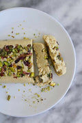 Slice of pistachio halva on a white plate, with additional pieces beside it, highlighting the ingredients and texture of the confection.