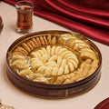 A beautifully arranged assortment of Zalatimo Sweets in a round gold tin, featuring various types of Middle Eastern cookies, including sesame cookies and buttery biscuits, displayed elegantly on a table with a traditional beverage in the background.