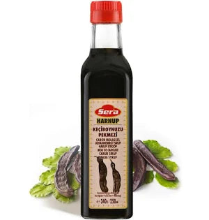 Bottle of Sera Carob Molasses (Harnup), showcasing a dark liquid with a red cap, placed against a background of carob pods and green leaves, highlighting its natural ingredients.